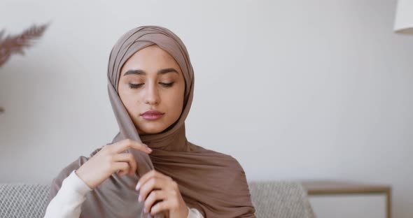 Islamic Traditional Headwear. Young Muslim Woman Putting on Hijab, Sitting on Couch at Home