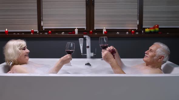 Senior Couple Grandfather and Grandmother Is Taking Foamy Bath, Drinking Red Wine in Bathroom