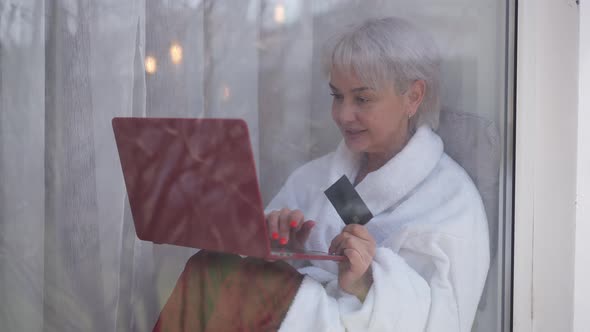 Joyful Mature Woman Sitting on Windowsill with Credit Card and Laptop Shopping Online