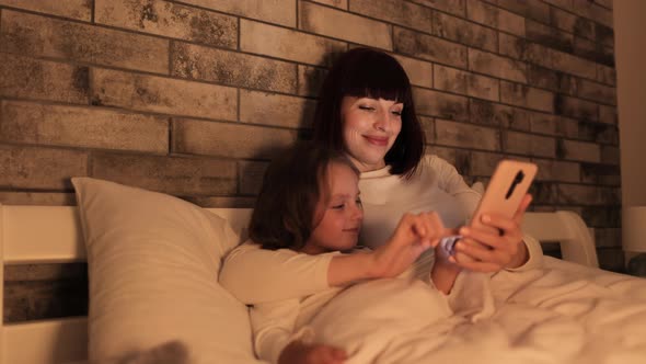 Mother and Cute Daughter Using Smartphone in the Evening Lying in Cozy Bed Covered with Blanket
