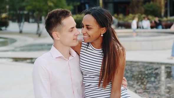 Close-up of Young Man Whispering Something in Girlfriend's Ear Sitting on a Bench Outdoors