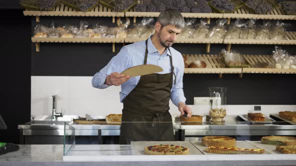 Young Handsome Man Vendor in the Apron Bringing Fresh Pies to the Counter in the Bakery Shop