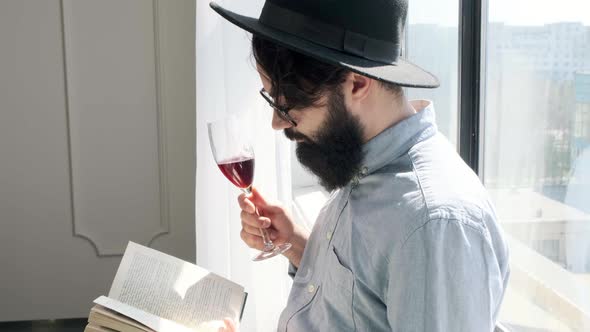 Pleased Bearded Man Drinking Red Wine While Reading a Book at Home