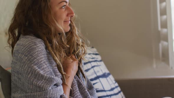 Caucasian woman sitting on sofa with blanket around shoulders smiling in sunny cottage living room