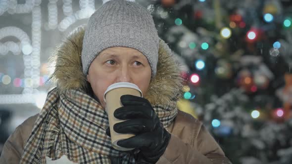 An Elderly Man Drinks Coffee From a Paper Cup in a Food Court at a Christmas Market in a Snowfall
