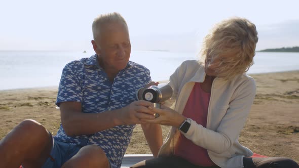 Happy Senior Couple Sitting on Plaid with Thermos Bottle on Summer Beach