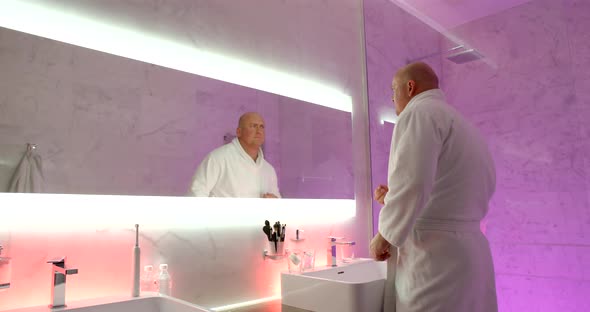 Adult Bald Man Is Wearing White Bathrobe Is Washing Face in Bathroom of Luxury Hotel in Morning Time