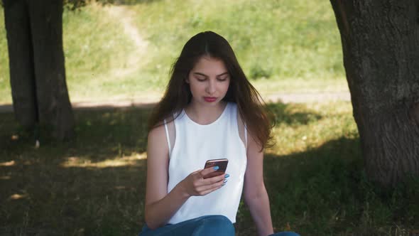 Beautiful Cheerful Girl with a Smartphone Sitting in a Park on a Bench on a Sunny Day Teenager