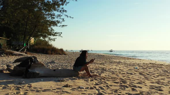 Young woman texting on her mobile phone, sitting on dried trunk over white sand of exotic beach with
