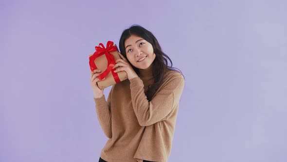 Asian Woman Smiling Holding Gift Box Tied with Red Ribbon Shaking It Trying to Guess What is There