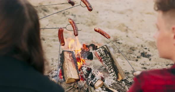 Close View of Friends Frying Sausages Sitting Around Bonfire on Sandy Beach