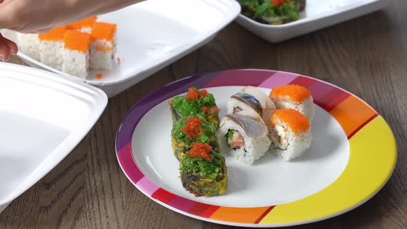 Woman Hand is Holding Bamboo Sticks and Shifting Sushi on a Colorful Plate Out of Dish Container