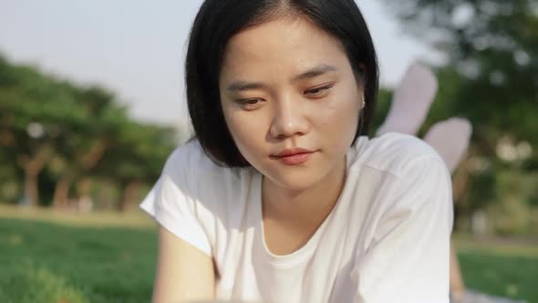 Asian female relaxed reading a book while lying on the floor grass outdoors at a public park.