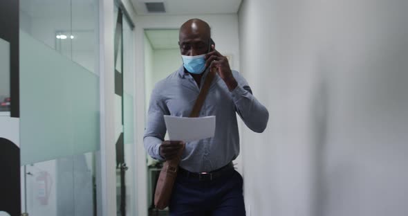 African american businessman in face mask holding document talking on smartphone in office corridor