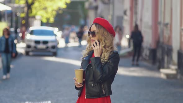 A Young Woman in the Middle of the Street is Talking on a Cell Phone and Drinking Takeaway Coffee