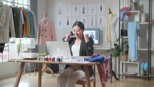 Asian Female Designer In Business Suit Working On A Laptop And Celebrating While Designing Clothes