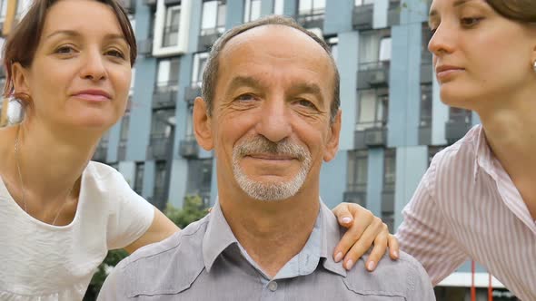 Family Portrait of Senior Father and Two Adult Daughters Kissing Him Outdoors on Modern Building of