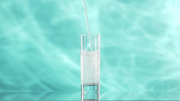 Foamy Clear Water is Pouring Into Collins Glass Standing on Cyan Smoky Background in Slowmotion