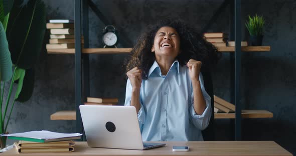 Excited Young African American Business Woman Enjoying Winning Sits at Table with Laptop Computer