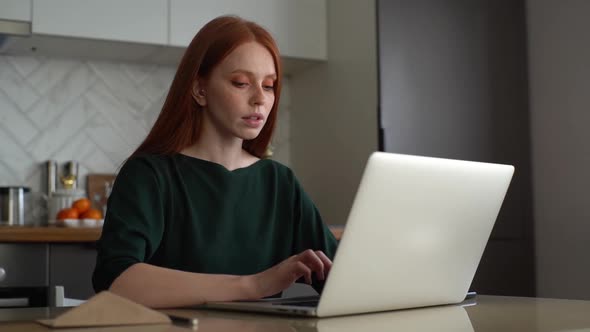 Focused Female Freelancer Working Typing on Laptop Computer Sitting at Table in Kitchen Room with