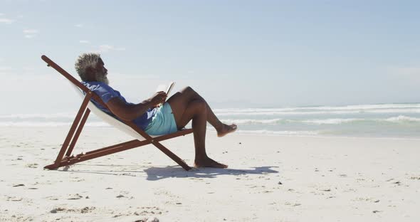 Senior african american man reading and lying on sunbed on sunny beach