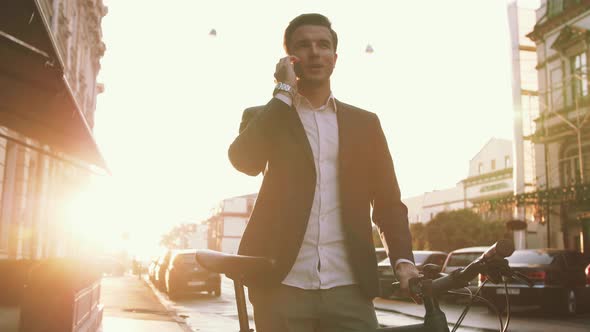 Handsome Young Stylish Man Talking on Mobile Phone Outdoors with Bicycle in City Center During