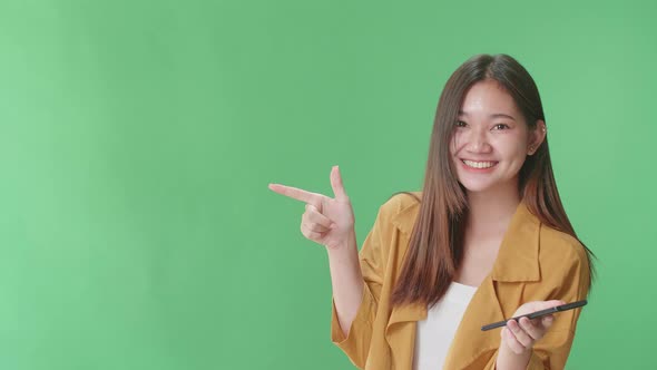 Asian Woman Use Phone With A Smile Pointing To The Side While Standing In The Green Screen Studio