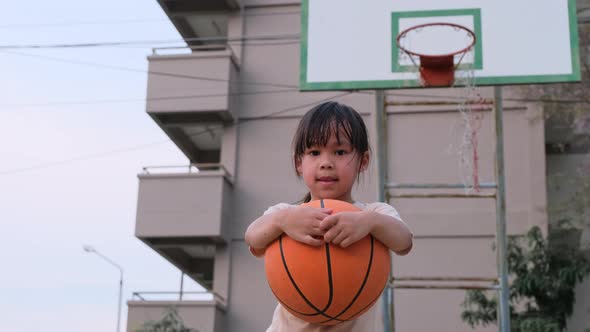 Cheerful cute girl holding basketball looking at camera at outdoor basketball playground. alt
