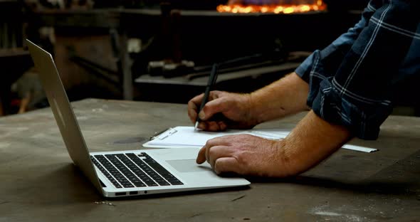 Blacksmith writing on clipboard while using laptop in workshop 4k