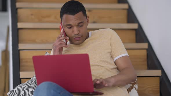 Middle Shot Front View of Thoughtful African American Man Talking on the Phone Typing on Laptop