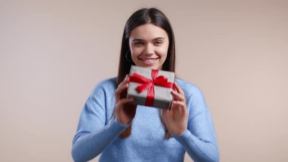 Woman Gives Gift Box By Hands to Camera on Light Wall Background