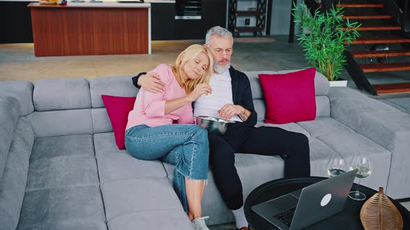 Older Man with Good Looking Blonde Watching Laptop on Coffee Table and Two Wine Glasses Beside