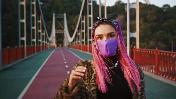 Young Hipster Woman in Informal Outfit and Protective Mask is Standing on Bridge Touching Her Pink