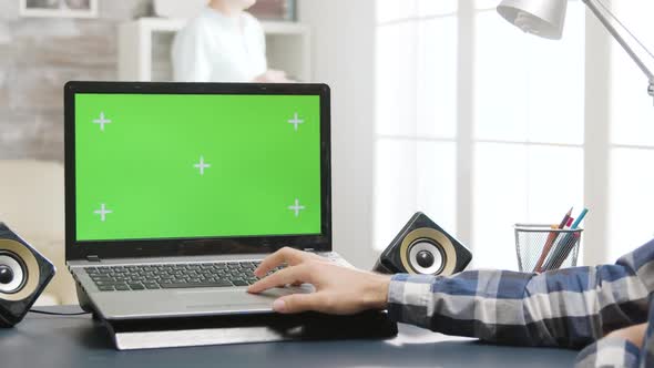 Man Sitting at the Desk in Bright and Well Lit Living Room Looking at Laptop with Green Screen