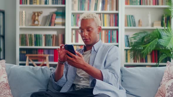 Smiling African Man Sits on Comfortable Sofa in Living Room Uses Mobile Phone