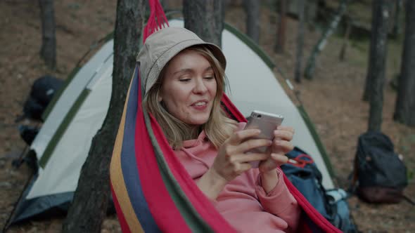 Joyful Young Woman Using Smartphone Texting and Laughing Over Messages Relaxing in Hammock in Forest
