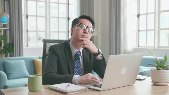 Asian Businessman Thinking And Writing In Notebook While Using Computer For Working At Home