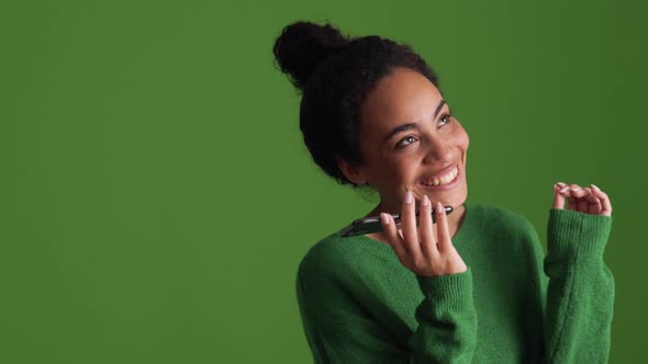 Smiling African woman in green shirt recording voice message by phone