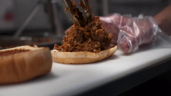 Cook uses tongs to place chopped beef on toasted bun, barbecue sandwich, close up slow motion 4K