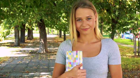 Young Pretty Blond Woman Holds Book and Smiles - Park with Trees in Background