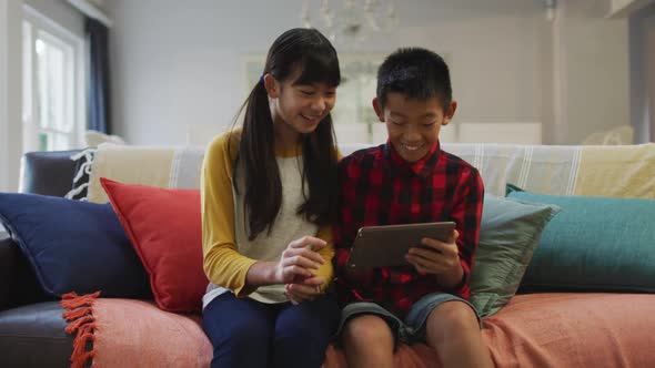 Asian brother and sister smiling and using tablet sitting on sofa at home