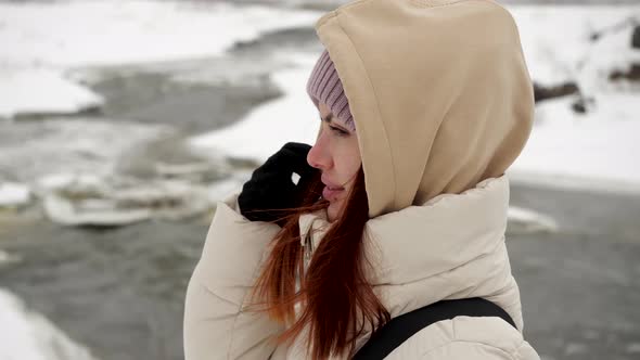 Attractive Girl Traveler in a Warm Jacket and Hood Stands in the Winter Forest