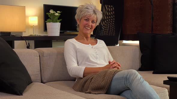 A Happy Middle-aged Woman Sits on a Couch in an Apartment Living Room and Applauds