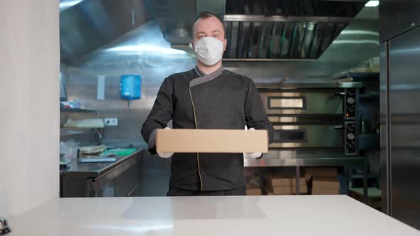 Portrait of Confident Caucasian Cook in Covid Face Mask Stretching Paper Box with Takeaway Order