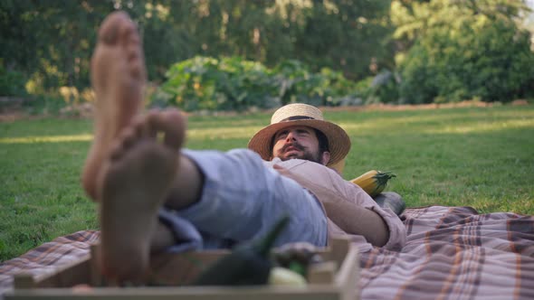 View From Feet of Young Bearded Farmer in Straw Hat Lying on Sunny Meadow Taking a Nap