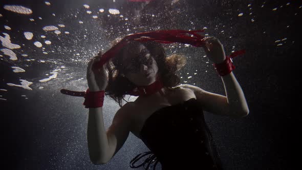 A Darkhaired Woman in Red Handcuffs and with a Red Whip Swims Deep Under the Water on a Dark