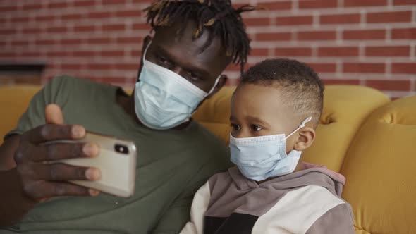 Father and Son are Sitting on a Sofa in Medical Face Masks Watching Video on Smartphone Close Up