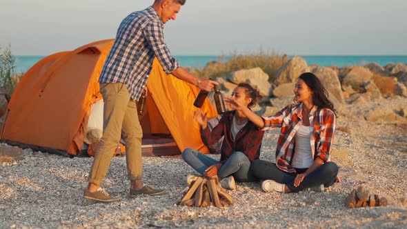 Young Adult Man in Casual Wear Brings Bottles of Beer To Girlfriends Sitting at Tent on Beach