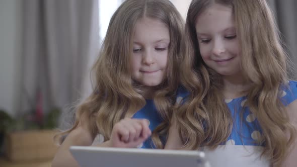 Close-up of Two Pretty Caucasian Twin Sisters with Long Wavy Hair Using Tablet. Portrait of Cute