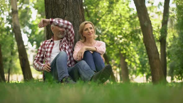 Optimistic Retired Couple Relaxing on Grass in Park, Planning Summer Weekend